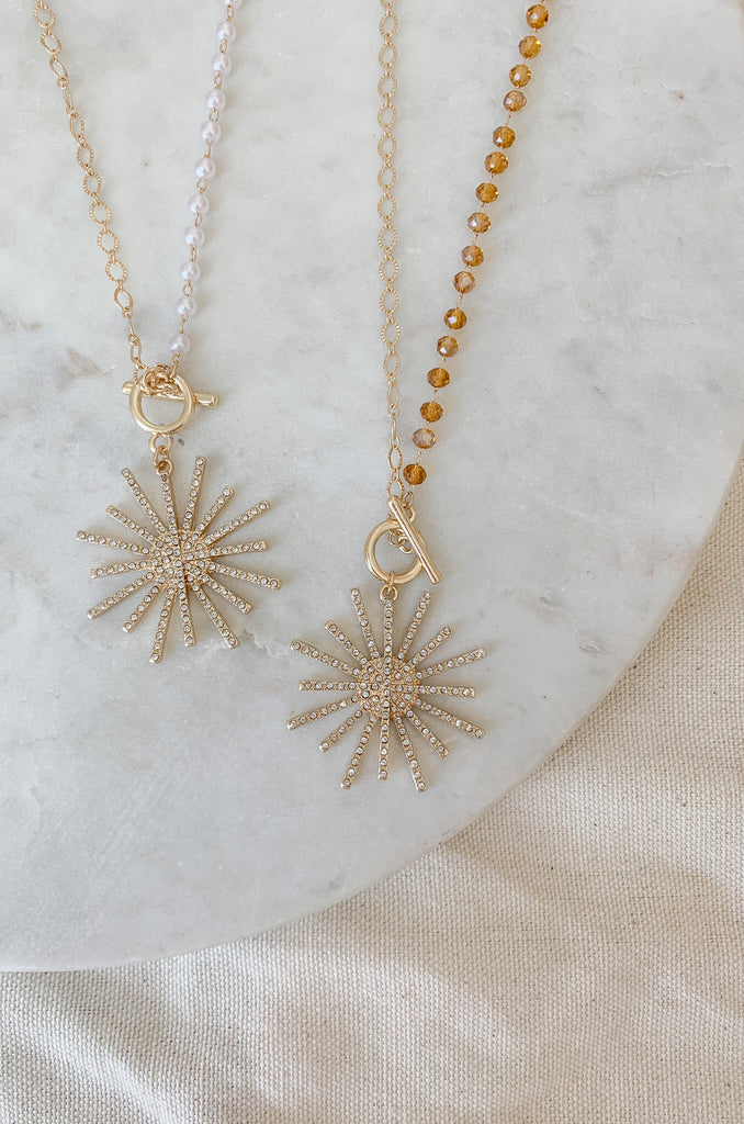 Light up your look with this ultra-dazzling Bead & Chain Toggle Starburst Necklace! This statement piece will sparkle around your neck and make you the star of any show. Choose between two golden looks with either pearl or topaz colored beads.