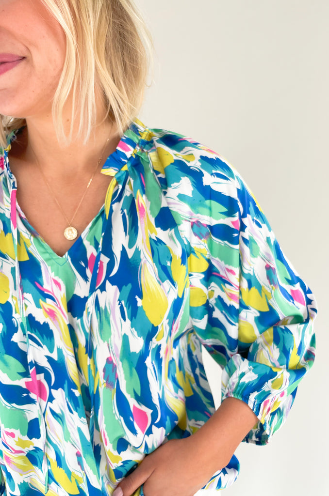 Long sleeve blouse with pops of vibrant blues, yellow, and pinks, flowy with balloon sleeve, lightweight silky feel, V-neck with a tie detailing and pleated neckline