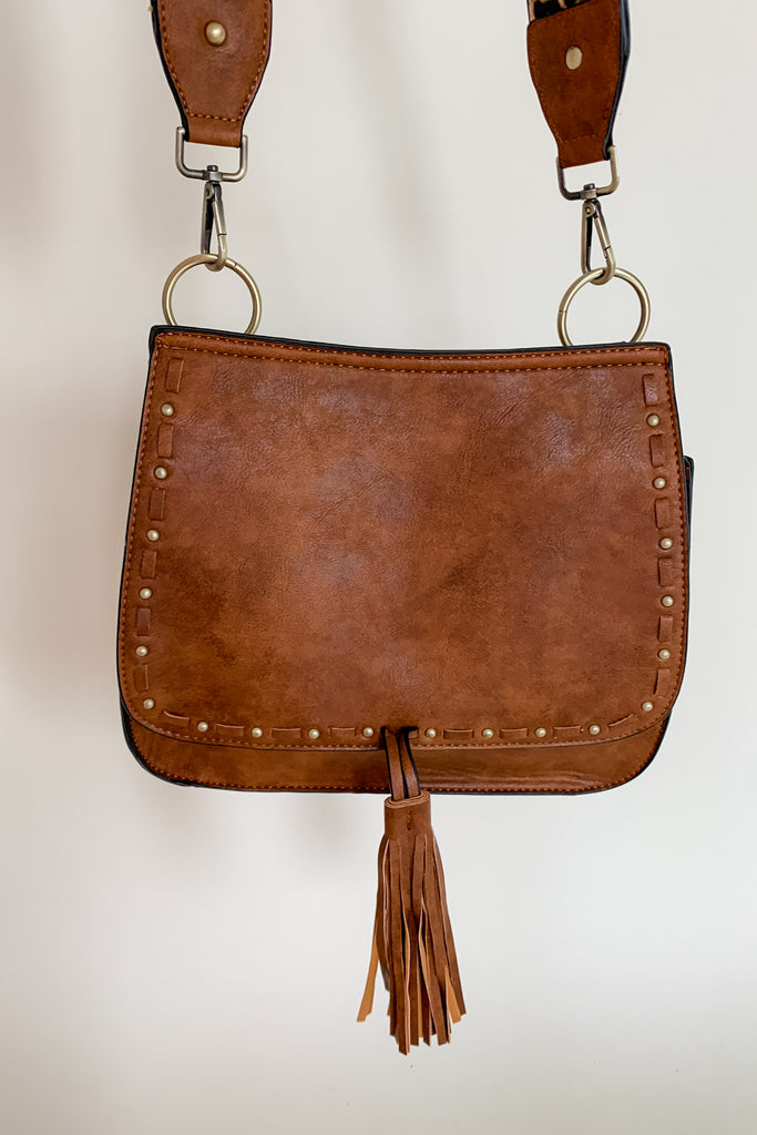 The Baylor Flapover Tassel Crossbody comes in three great colors: Brown, Teal, and Olive. It's a square shape that tapers at the top. Because of the unique shape, it fits comfortably right at the hip. Each bag comes with an exclusive guitar strap. On the flap, there are hammered metallic dots that embroider it. There is also a unique woven stitching followed by a tassel. This bag is right on trend this season, the perfect size, and all vegan leather! 