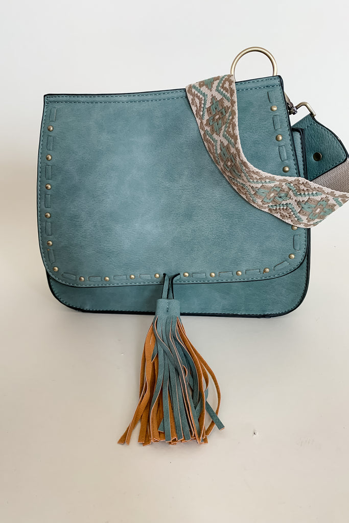 The Baylor Flapover Tassel Crossbody comes in three great colors: Brown, Teal, and Olive. It's a square shape that tapers at the top. Because of the unique shape, it fits comfortably right at the hip. Each bag comes with an exclusive guitar strap. On the flap, there are hammered metallic dots that embroider it. There is also a unique woven stitching followed by a tassel. This bag is right on trend this season, the perfect size, and all vegan leather! 