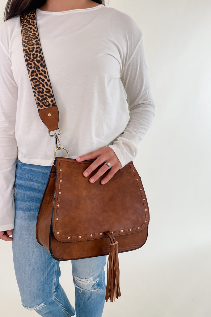 The Baylor Flapover Tassel Crossbody comes in three great colors: Brown, Teal, and Olive. It's a square shape that tapers at the top. Because of the unique shape, it fits comfortably right at the hip. Each bag comes with an exclusive guitar strap. On the flap, there are hammered metallic dots that embroider it. There is also a unique woven stitching followed by a tassel. This bag is right on trend this season, the perfect size, and all vegan leather! 