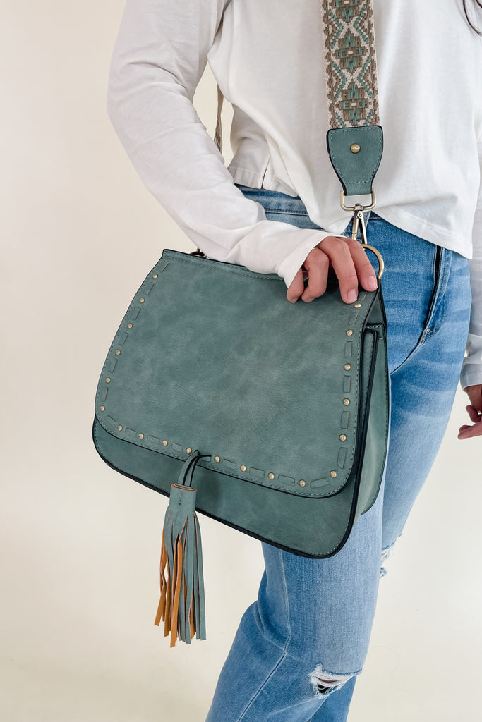 The Baylor Flapover Tassel Crossbody comes in three great colors: Brown, Teal, and Olive. It's a square shape that tapers at the top. Because of the unique shape, it fits comfortably right at the hip. Each bag comes with an exclusive guitar strap. On the flap, there are hammered metallic dots that embroider it. There is also a unique woven stitching followed by a tassel. This bag is right on trend this season, the perfect size, and all vegan leather! 